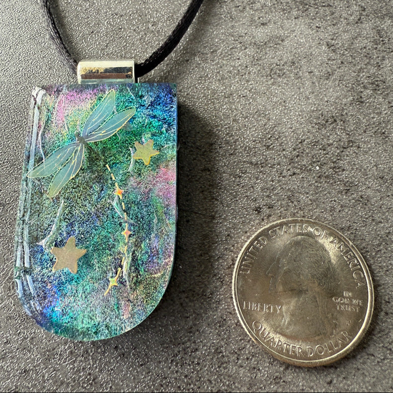 Colorful pendant with dragonflies and stars next to a quarter on a gray surface