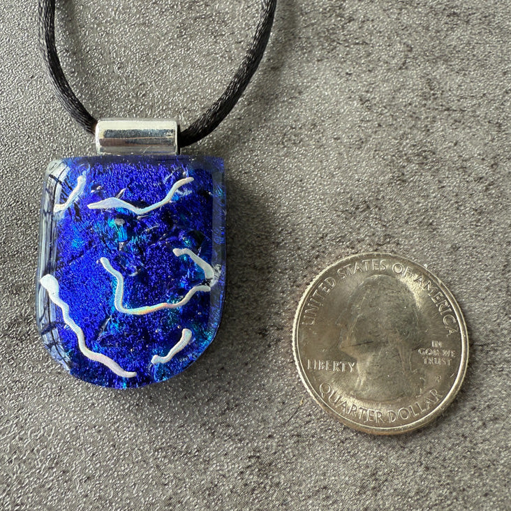 Blue pendant with white designs on a gray surface next to a penny