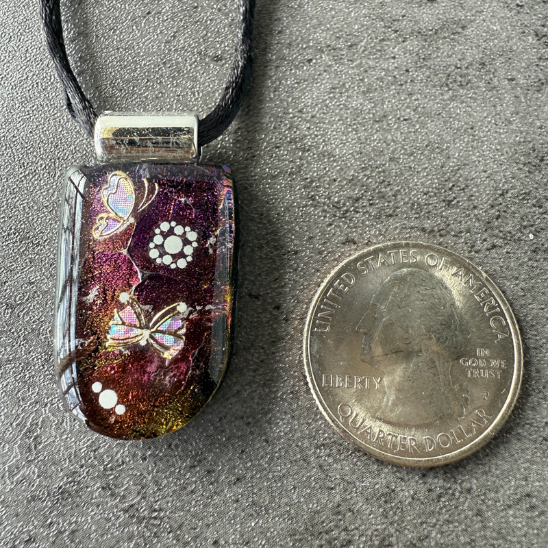 Decorative pendant with butterfly designs next to a U.S. quarter dollar coin on a gray surface.